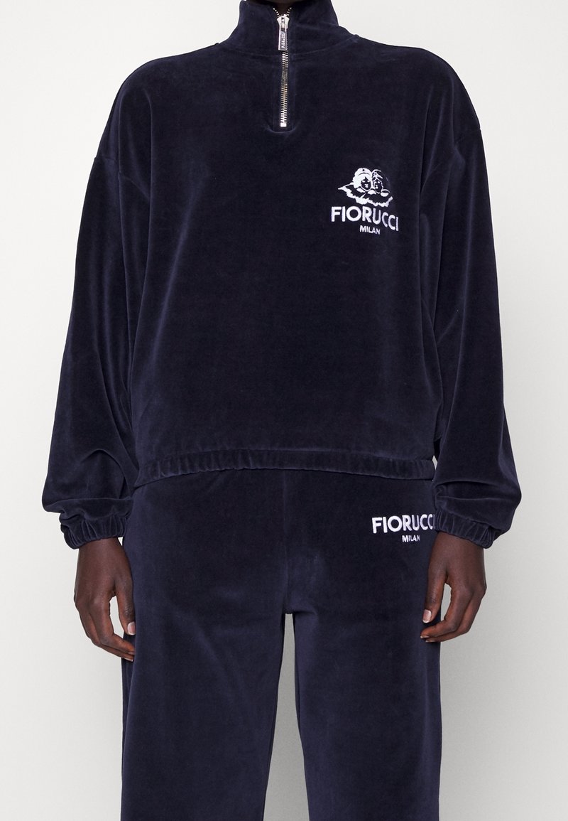 Survêtement en velours bleu marine avec un pull à demi-fermeture éclair et un pantalon assorti, les deux arborant les logos blancs Fiorucci Milan sur la poitrine et la cuisse.