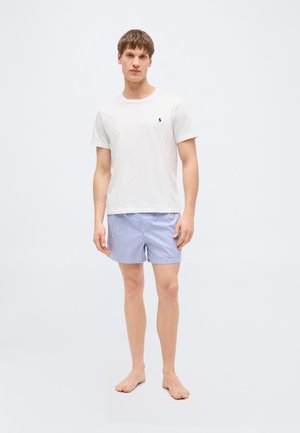 Fiatal férfi mezítláb áll, fehér rövid ujjú ingben és kék-fehér csíkos rövidnadrágban egy egyszínű háttér előtt.