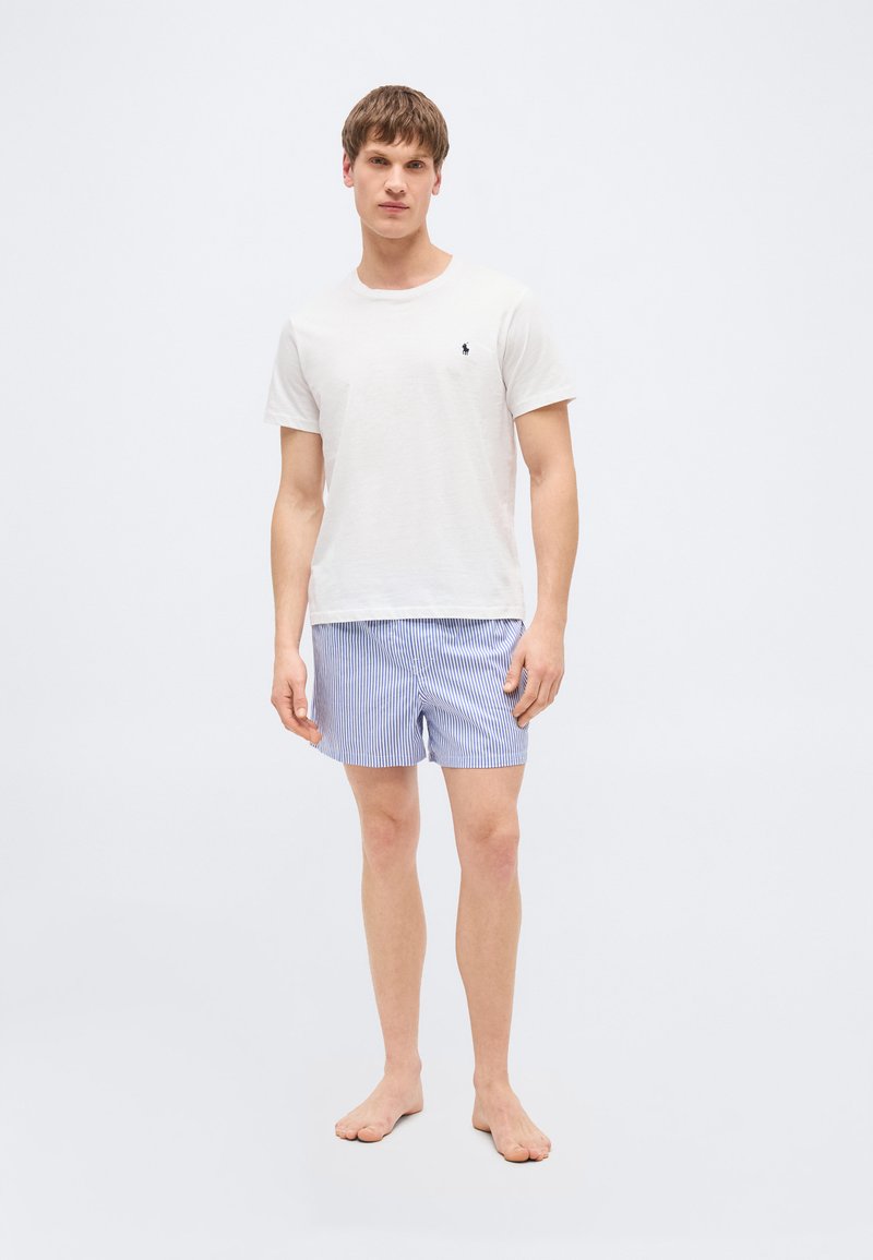 Νέος άνδρας που στέκεται ξυπόλητος, φοράει λευκό πουκάμισο με κοντά μανίκια και μπλε-άσπρες ριγέ βερμούδες, μπροστά σε απλό φόντο.