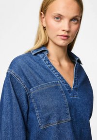 Tumši zils džinsu krekls ar V veida kaklu un vienu krūšu kabatu. Iezīmējas ar brīvu piegriezumu, redzamām šuvēm un mīkstu tekstūru.