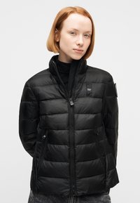 Jeune femme aux cheveux roux courts portant une veste matelassée noire avec des poches zippées et se tenant devant un fond blanc.