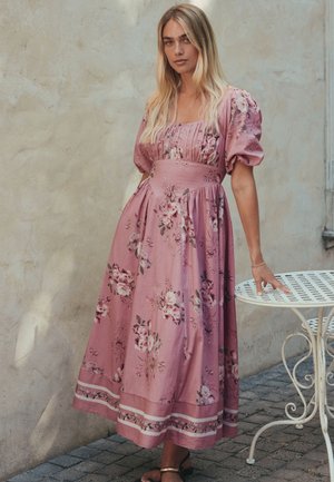 Frau in einem rosa Midikleid mit Blumenmuster und Puffärmeln, die neben einem weißen schmiedeeisernen Tisch vor einer beigen Wand steht.