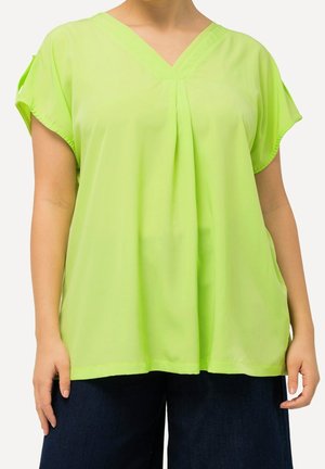 Femme portant un chemisier ample vert citron à manches courtes avec un col en V et un détail plissé, associé à un pantalon large foncé.