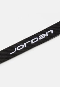 Juodas audinys diržas su baltu "Jordan" tekstu. Diržas turi tekstūruotą paviršių su smulkiai audžiomis detalėmis.