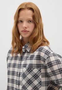Camicia a quadri nei toni crema, blu e nero. Presenta un colletto con bottoni e una tasca sul petto con motivo a quadri. Tessuto morbido.