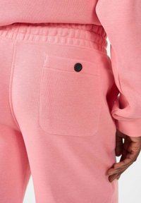 Roze joggingbroek met een zachte textuur, een elastische tailleband en een achterzak met een zwarte knoopdetail. Gericht op comfort en casual stijl.
