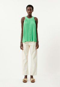 Groene mouwloze top met een geplooide halslijn en knoopdetail, gecombineerd met crèmekleurige broeken en beige plateau sandalen.