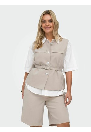 Žena v béžovej veste so striebornými gombíkmi a opaskom, cez bielu košeľu, a béžových krátkych nohaviciach do kolien, usmieva sa s uvoľnenými rukami.