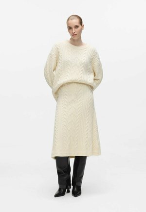 Kvinde iført en cremefarvet kabelstrikket sweater og matchende nederdel med sorte hælede støvler, stående mod en hvid baggrund.
