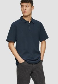 Navy blue polo shirt with short sleeves, collared neck, and three golden buttons. Worn with black textured pants.