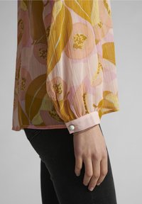 Langärmlige Bluse aus leichtem, plissiertem Stoff. Sie hat ein florales Muster in Gelb und Pink, mit einem gerafften Ärmel im Seidenstil und einem Knopfdetail.