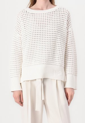Femme portant un pull en maille blanche ajourée à manches longues avec un ourlet côtelé, associé à un pantalon blanc ample avec une ceinture en tissu.