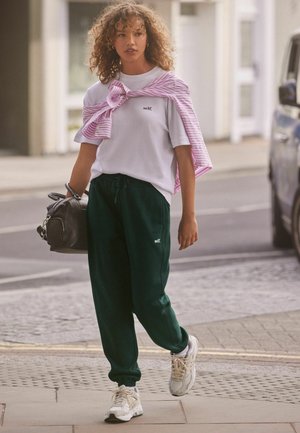 White crew-neck t-shirt with "self." logo, green sweatpants, pink striped sweater draped over shoulders, carrying black handbag, wearing white sneakers.