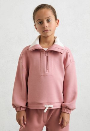 Noor tüdruk, kellel on taga seotud juuksed, kannab roosat lukuga dressipluusi ja sobivaid pükse heleda tausta ees.