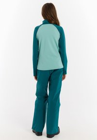 Turquoise softshelljack met hoge kraag en lange mouwen, gecombineerd met bijpassende broeken. De outfit heeft een soepele textuur en een relaxte pasvorm.