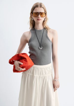 Femme portant des lunettes de soleil ambrées, un haut gris sans manches, une jupe crème, des créoles et un collier, tenant un sac rouge sur fond uni.