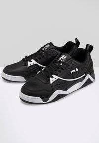 Paire de baskets FILA noires et blanches avec des semelles épaisses, un design à lacets et le logo de la marque sur le côté et la languette, sur un fond gris et blanc.