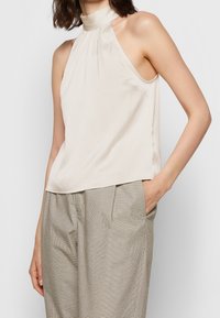 Blouse sans manches beige avec un col montant et un tissu texturé, assortie à un pantalon à carreaux gris. La blouse présente une coupe ample et un léger drapé.