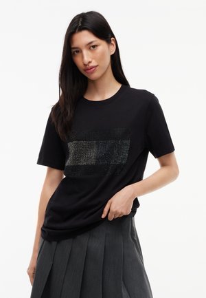 Jeune femme aux longs cheveux foncés porte un t-shirt noir avec un motif rectangulaire scintillant et une jupe plissée gris foncé.