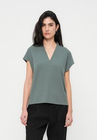 Κοντομάνικη μπλούζα με V λαιμόκοψη σε απαλό πράσινο ύφασμα, με δομημένη εφαρμογή και απαλή υφή, σε συνδυασμό με μαύρο, άνετο παντελόνι.