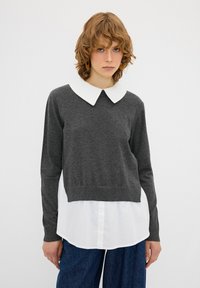 Grauer Pullover mit langen Ärmeln und einem weißen Hemd darunter. Der Pullover hat einen cropped Schnitt und eine weiche Textur. Denim-Hosen runden das Outfit ab.