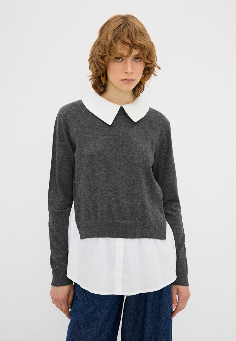 Grauer Pullover mit langen Ärmeln und einem weißen Hemd darunter. Der Pullover hat einen cropped Schnitt und eine weiche Textur. Denim-Hosen runden das Outfit ab.