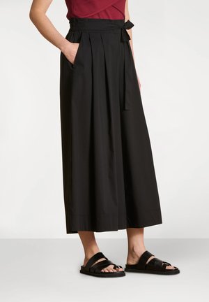 Pantalon large taille haute noir avec plis, poches latérales et ceinture nouée. Porté avec des sandales slides noires sur un fond clair.