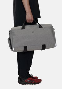 Grå tyg resväska med svarta handtag och detaljer, med en rund logotyp och en sidofack med dragkedja. Rektangulär form med robust design.