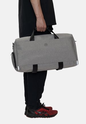Grå stof rejsetaske med sorte håndtag og detaljer, der har et rundt logo og en sidelomme med lynlås. Rektangulær form med robust design.