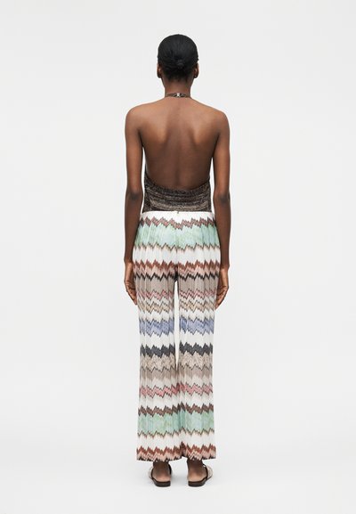 Γυναίκα με σκουρόχρωμο δέρμα και μαλλιά σε κότσο, φορώντας ένα τοπάκι χωρίς πλάτη και παντελόνες με σχέδιο ζιγκ ζαγκ, ψηλόμεσες και με φαρδιά πόδια, στέκεται με την πλάτη προς τα έξω.