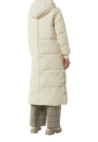Langer cremefarbener Daunenmantel mit Kapuze, mit horizontalem gesteppte Muster, seitliche Schlitze für Bewegungsfreiheit, getragen über karierter Hose und klobigen Boots.