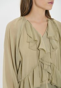 Blusa verde oliva chiaro realizzata in tessuto trasparente, con volant lungo il collo e le maniche, caratterizzata da una silhouette fluida e una texture morbida.