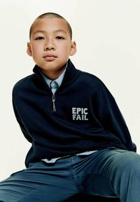 Chlapec s krátkymi vlasmi sedí sebavedome, oblečený v námorníckej zipsovej mikine s nápisom "EPIC FAIL", pod ktorou má na sebe košeľu s golierom.