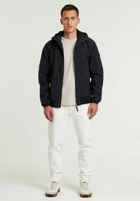 Chaqueta exterior navy con cierre de cremallera, bolsillos frontales, capucha ajustable y puños de canalé. Llevada con unos jeans claros y zapatillas deportivas.