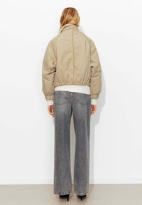 Veste courte beige à col haut et taille élastique, associée à un pantalon en denim gris ample et des talons noirs.