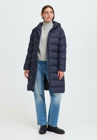 Long navy puffer coat with a hood, quilted design, front zipper, and side pockets. Worn over a grey knitted sweater and blue jeans.