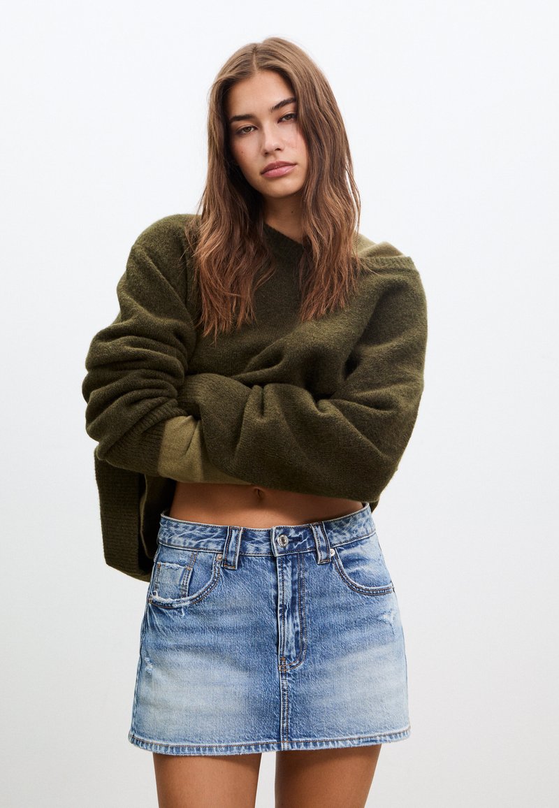 Pull court vert olive en tissu texturé ; mini-jupe en jean bleu clair avec cinq poches et zones décolorées.