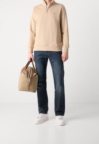 Sudadera beige con cremallera, vaqueros azul oscuro, zapatillas blancas y un bolso de lona marrón claro con detalles en marrón claro y cremallera.