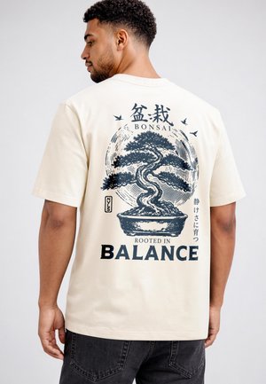 Man wearing cream t-shirt with black bonsai tree graphic, birds, and text "BONSAI" and "Rooted in Balance" on the back.