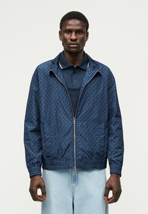 Mladý muž nosiaci námornícku modrú vzorovanú bundu na zips cez tmavomodré polotričko a svetlomodré rifle, stojaci na neutrálnej pozadí.