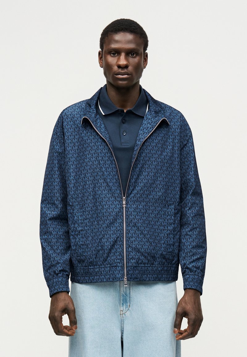 Νέος άνδρας που φορά μια μπλε σκούρα ζακέτα με φερμουάρ και σχέδια πάνω από ένα μπλε σκούρο πόλο πουκάμισο και ανοιχτόχρωμα μπλε τζιν, στέκεται μπροστά σε ουδέτερο φόντο.