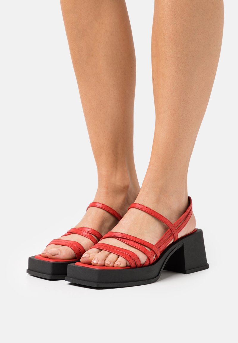 Pieds portant des sandales rouges à brides, avec des bouts carrés et des talons blocs noirs épais sur fond blanc.