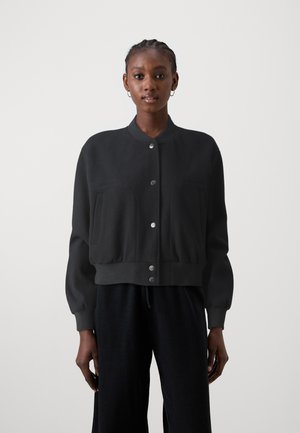 Chaqueta bomber corta negra de tejido suave, con cuello alzado, botones a presión y dos bolsillos frontales con bordes redondeados.