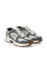Pár šedobílých běžeckých bot New Balance s síťovanými panely a hnědými gumovými podrážkami na bílém pozadí.