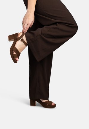 Persoon met bruine broek met wijde pijpen stelt de enkelriem van bijpassende bruine sandalen met blokhak af tegen een witte achtergrond.