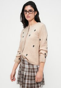 Μπεζ ζακέτα με λουλουδάτη κεντημένη διακόσμηση σε μπλε και πράσινο, υφαντό ύφασμα με υφή, στρογγυλή λαιμόκοψη και κούμπωμα με κουμπιά μπροστά, φορεμένη με καρό φούστα.