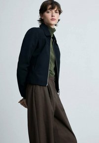 Giacca nera con zip e colletto, indossata sopra un dolcevita verde e pantaloni marroni a vita alta. Tessuto liscio, design su misura.