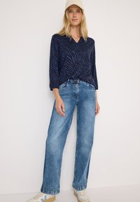 Haut marine avec des rayures zébrées bleues, décolleté en V et manches trois-quarts, associé à un jean large bleu clair et des baskets beiges.