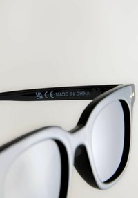Lunettes de soleil noires et grises avec des verres surdimensionnés, présentant une finition brillante, des accents dorés et un texte sur la branche indiquant les informations de fabrication.