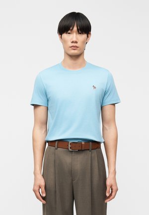 Mężczyzna o czarnych włosach nosi jasnoniebieski t-shirt z krótkim rękawem z małym logo zebry, brązowy skórzany pasek i wysokie spodnie w kolorze taupe, stoi prosto.